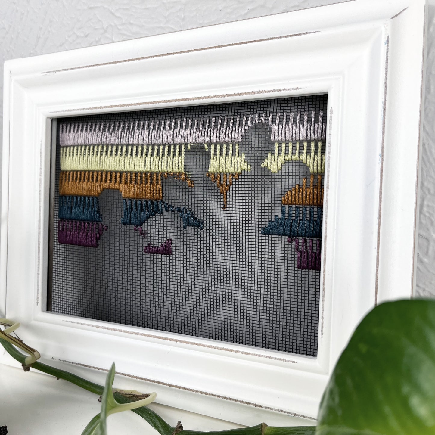 a close up angled view of a piece of window screen hand stitched with rows of lavender chartreuse brown midnight blue and plum stitches with an image in negative space of prickly pear cacti, in a white wood frame, on a white counter, with a pothos vine around it