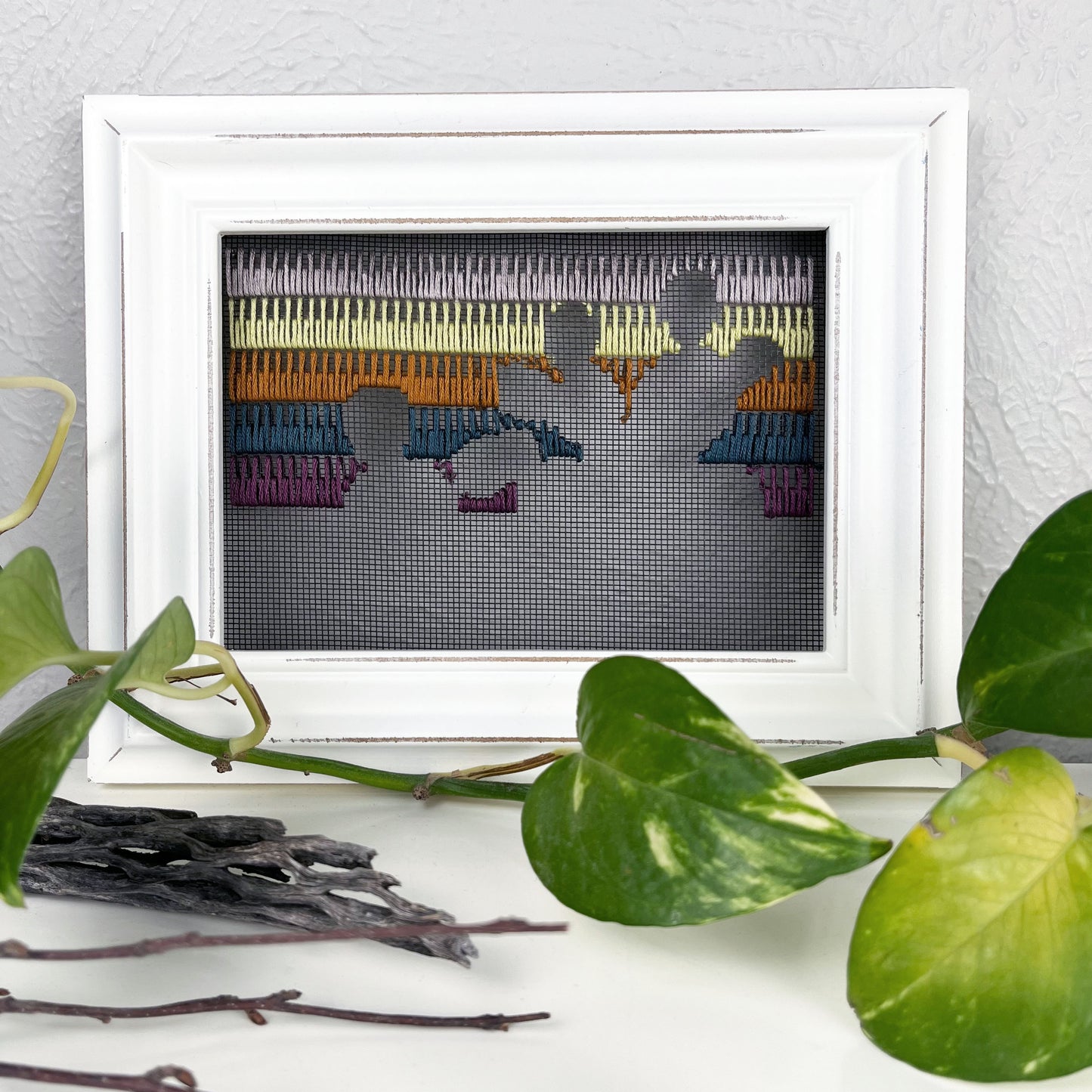 a piece of window screen hand stitched with rows of lavender chartreuse brown midnight blue and plum stitches with an image in negative space of prickly pear cacti, in a white wood frame, on a white counter, with a pothos vine around it