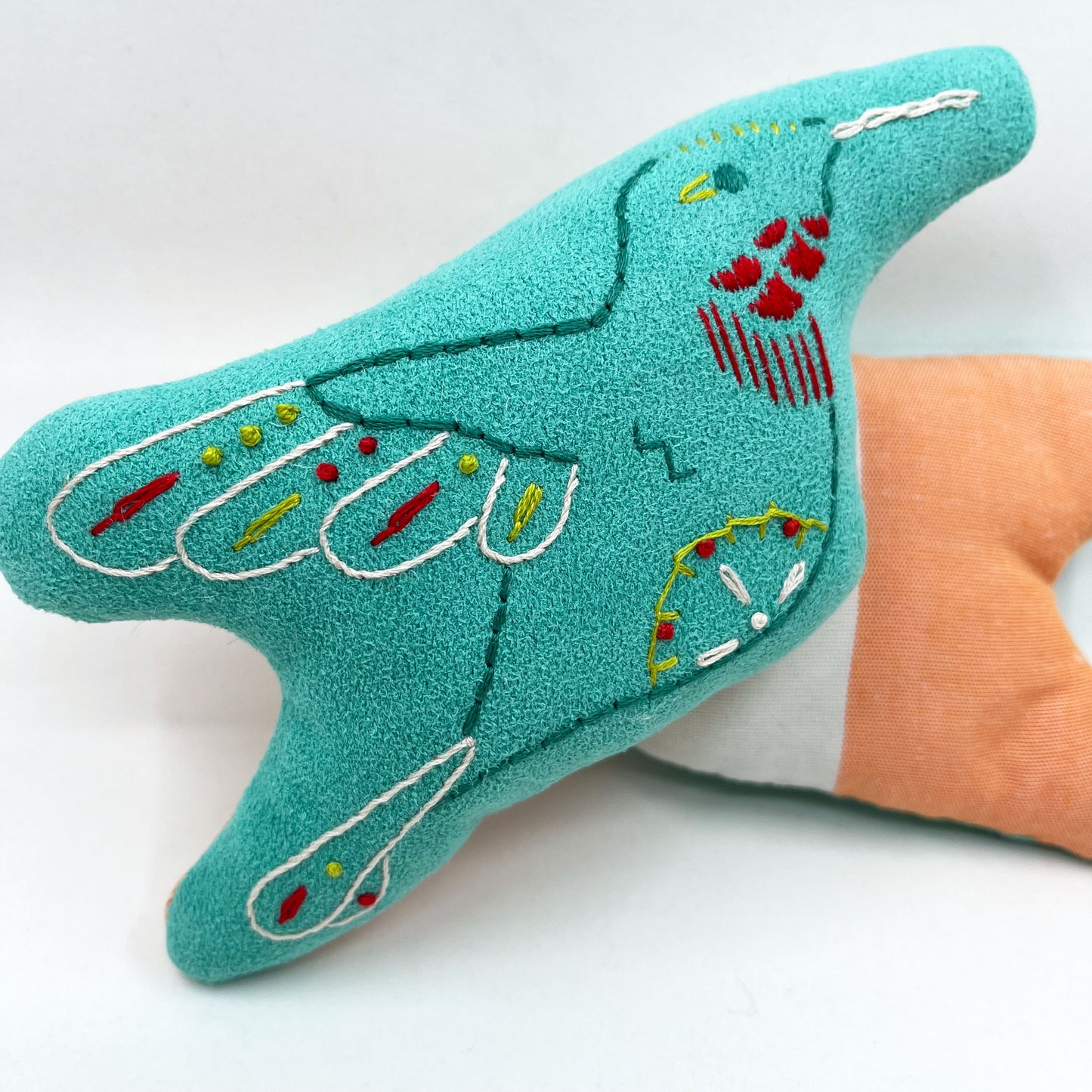 a colorfully hand embroidered stuffed pillow animal of a hummingbird on seafoam green fabric, behind it one turned around showing the back side made of half white half pink fabric