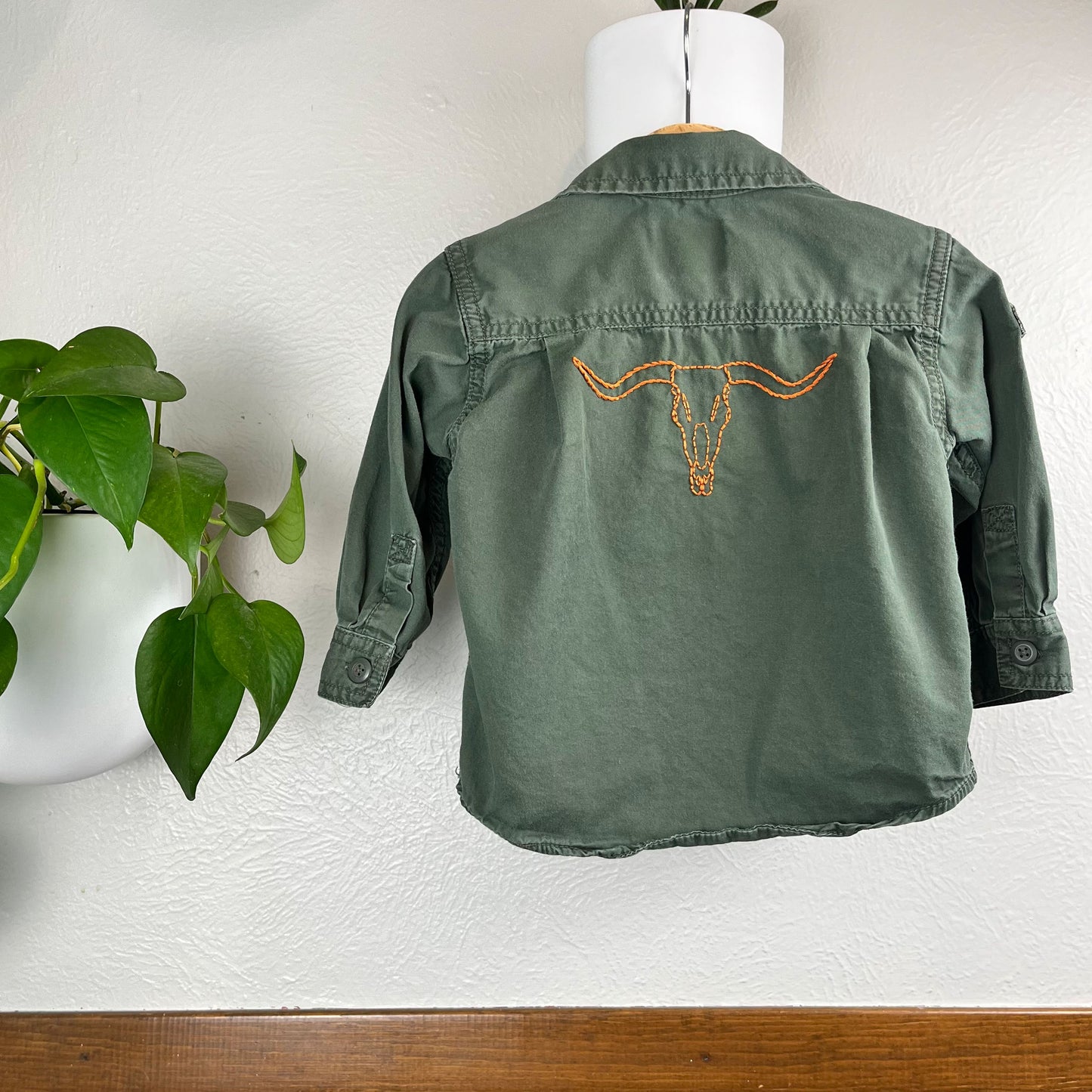 back view of a green long sleeved button up shirt hanging on a wall next to a pothos plant, with an outline of a longhorn skull embroidered in orange below the back yoke