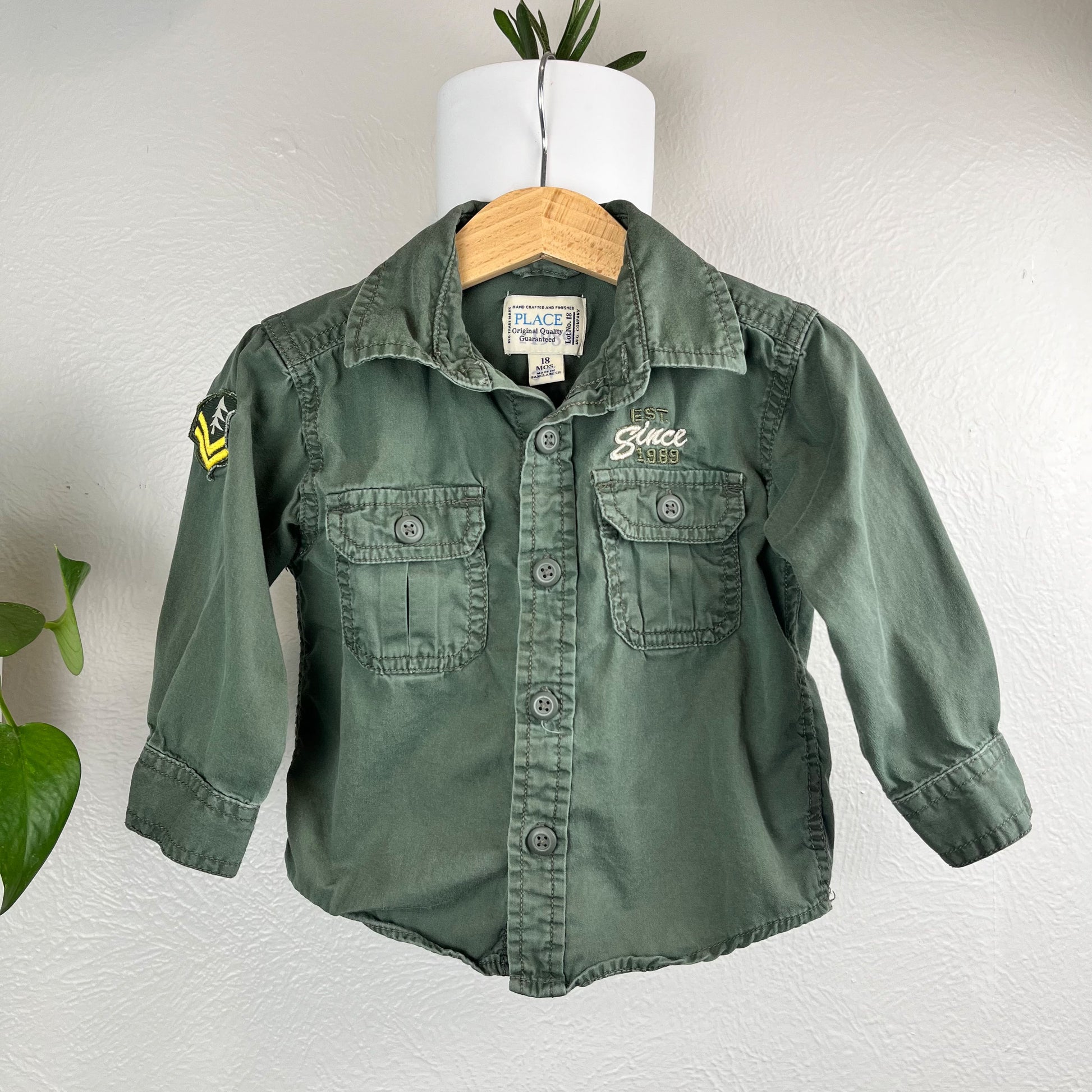 front view of a green long sleeved button up shirt hanging on a wall next to a pothos plant