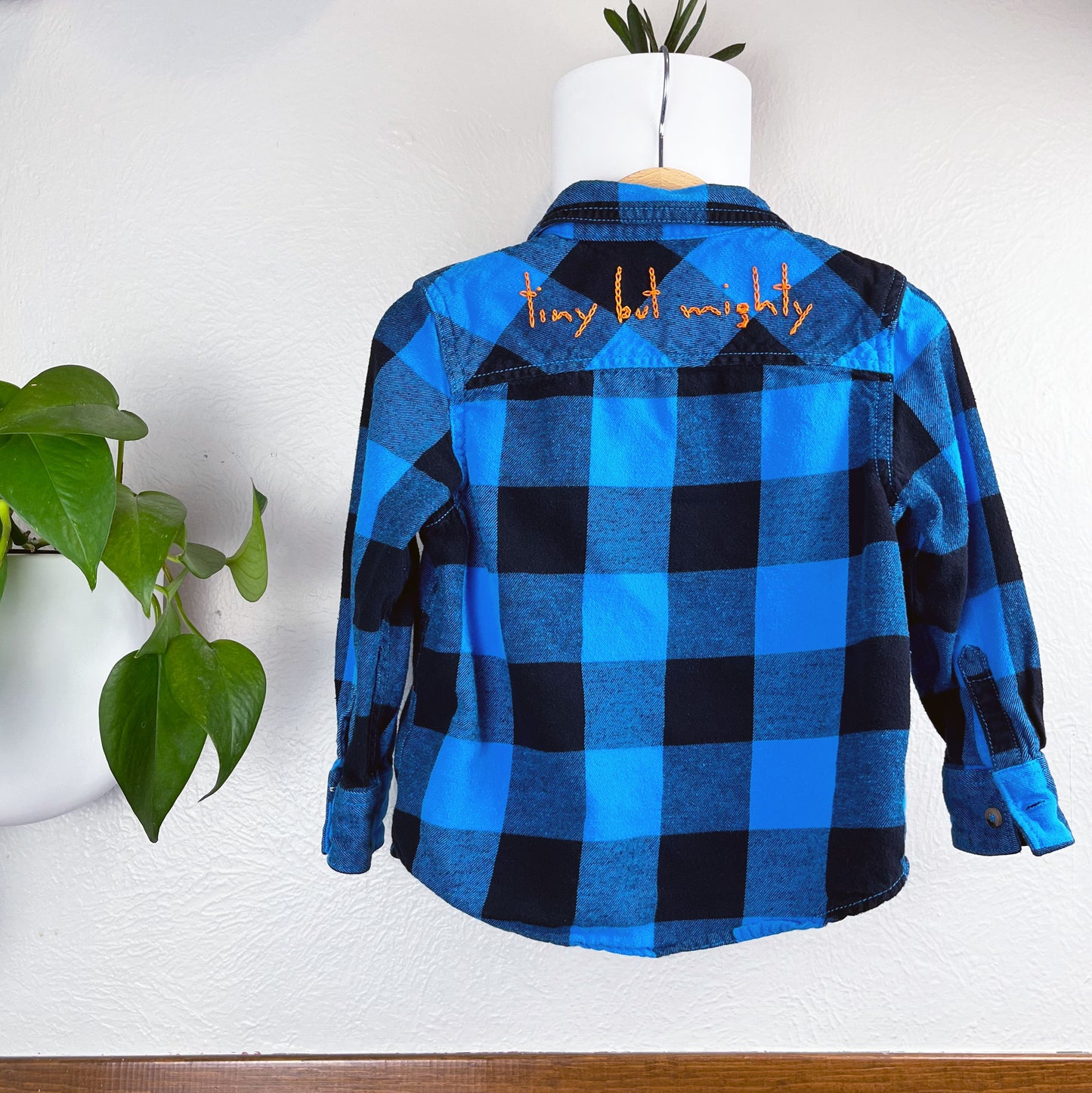 back view of a blue and black plaid flannel with the words "tiny but mighty" hand embroidered in a bright orange chainstitch on the back yoke, hanging on a wall next to a pothos plant 