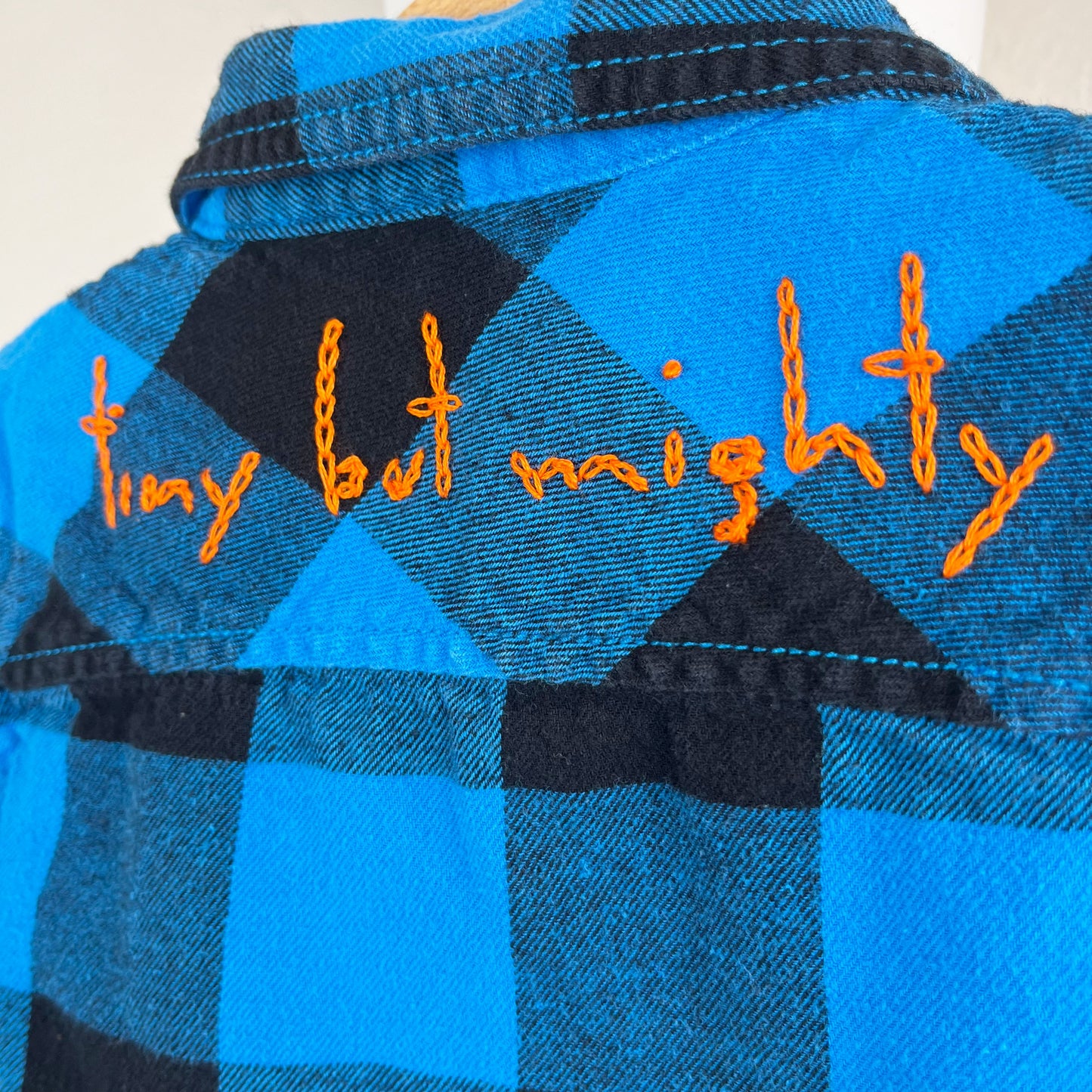 the words "tiny but mighty" hand embroidered in a bright orange chainstitch on the back yoke of a blue and black plaid flannel