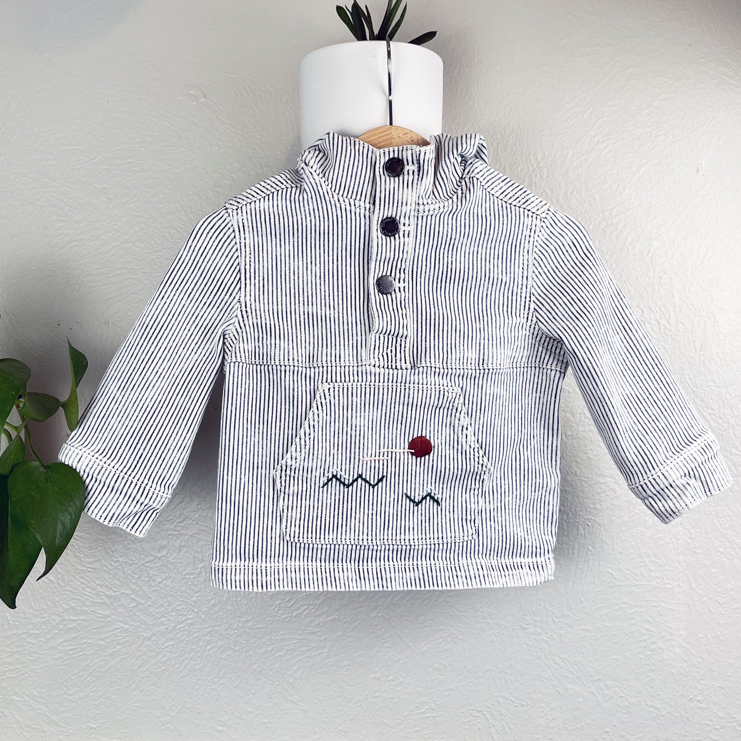 a railroad striped pullover with kangaroo pocket, with a hand embroidered abstract landscape on the pocket, hanging on a wall next to a pothos plant 