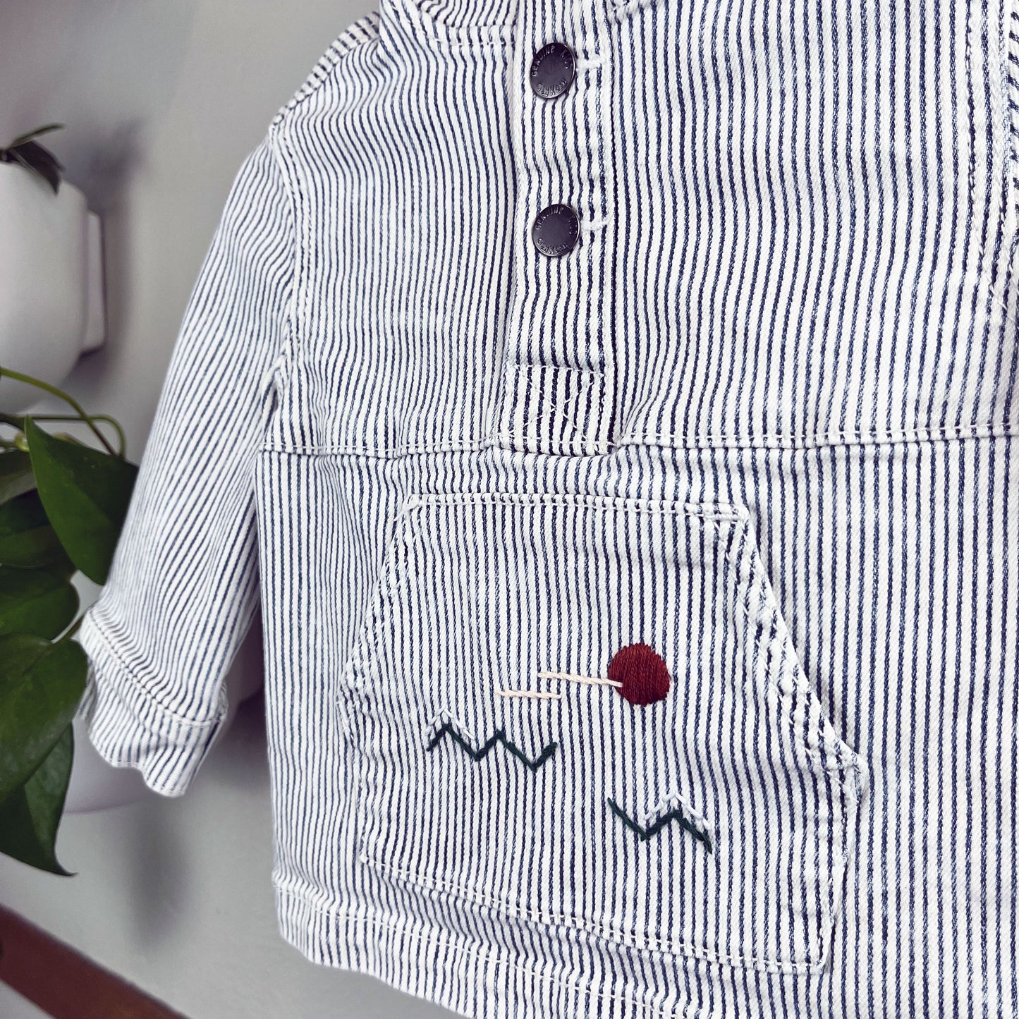 close up angled view of a railroad striped pullover with kangaroo pocket, with a hand embroidered abstract landscape on the pocket, hanging on a wall with a pothos plant in the background