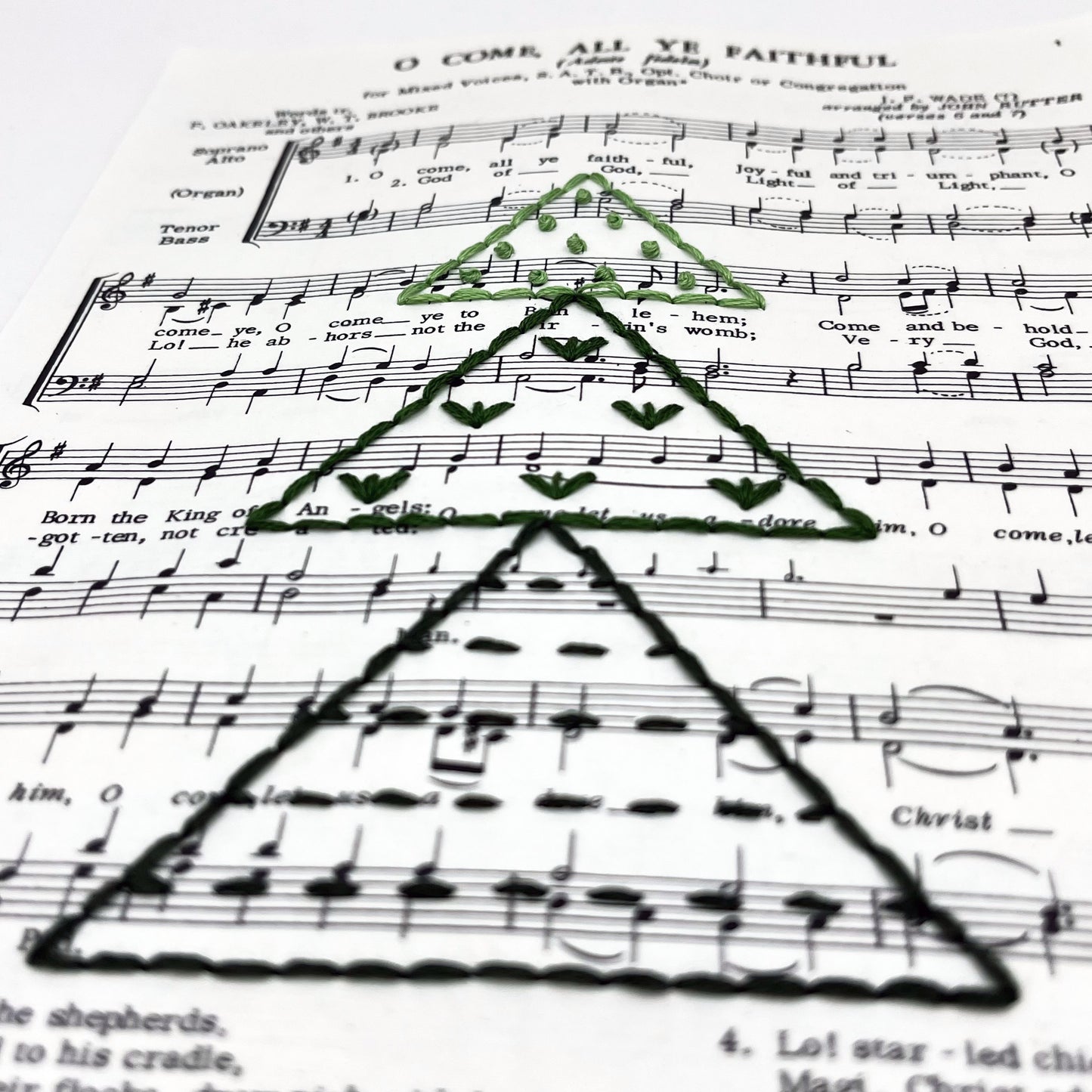 close up angled view of Christmas sheet music for the song O Come All Ye Faithful, hand stitched on with a Christmas tree made from triangles filled with different types of stitches, in shades of green thread