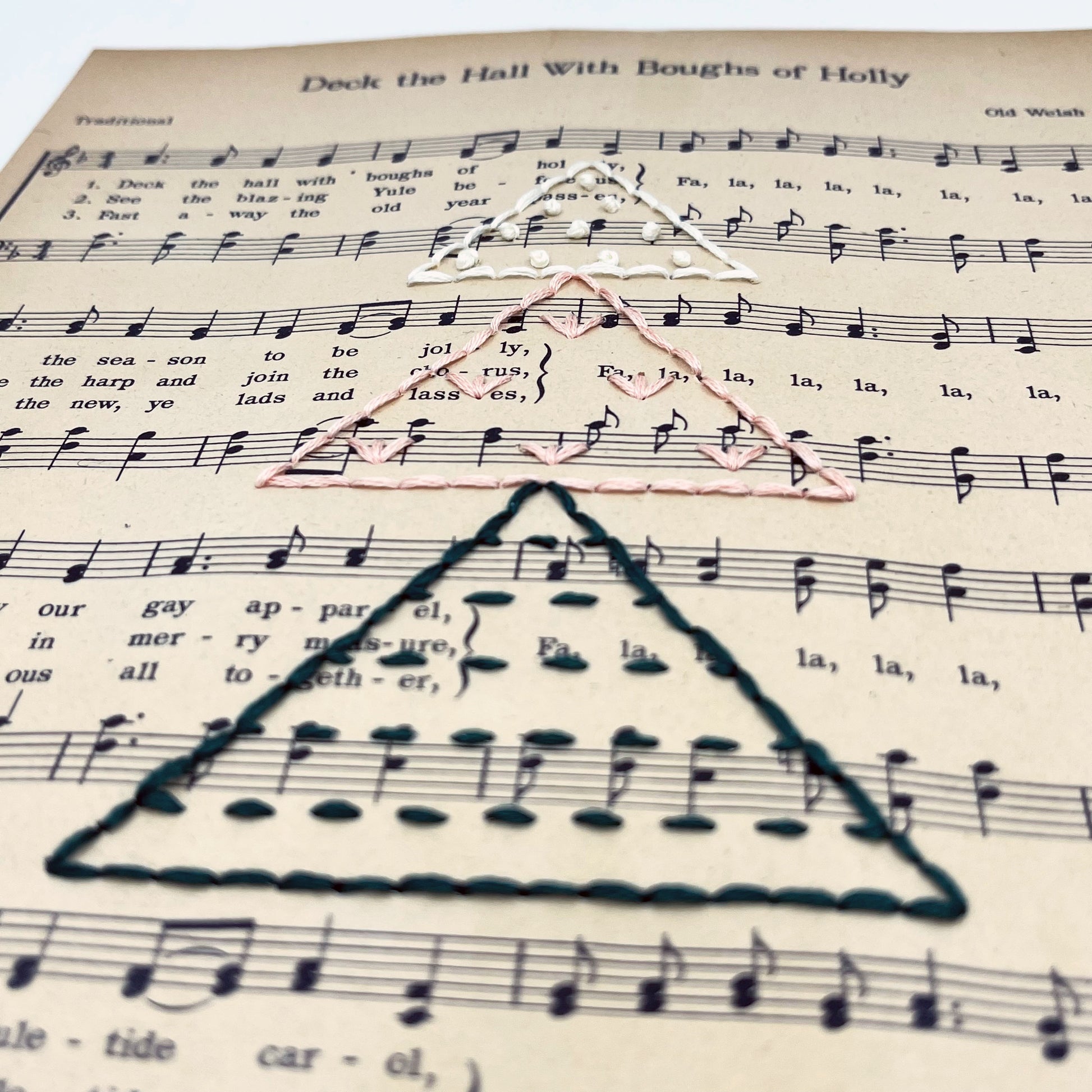 close up angled view of Christmas sheet music for the song Deck the Hall, hand stitched on with a Christmas tree made from triangles filled with different types of stitches, in green, peach and ivory thread