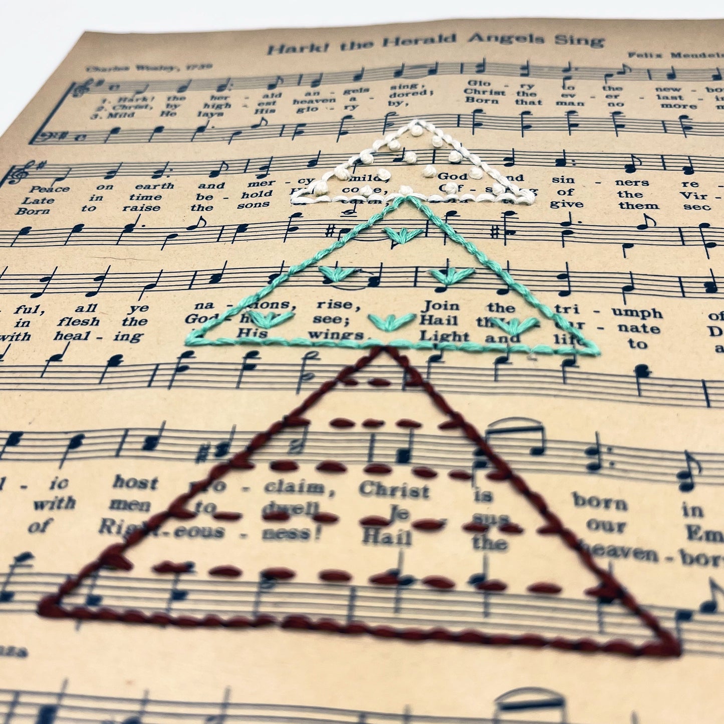 close up angled view of Christmas sheet music for the song Hark the Herald Angels Sing, hand stitched on with a Christmas tree made from triangles filled with different types of stitches, in red, green and ivory thread