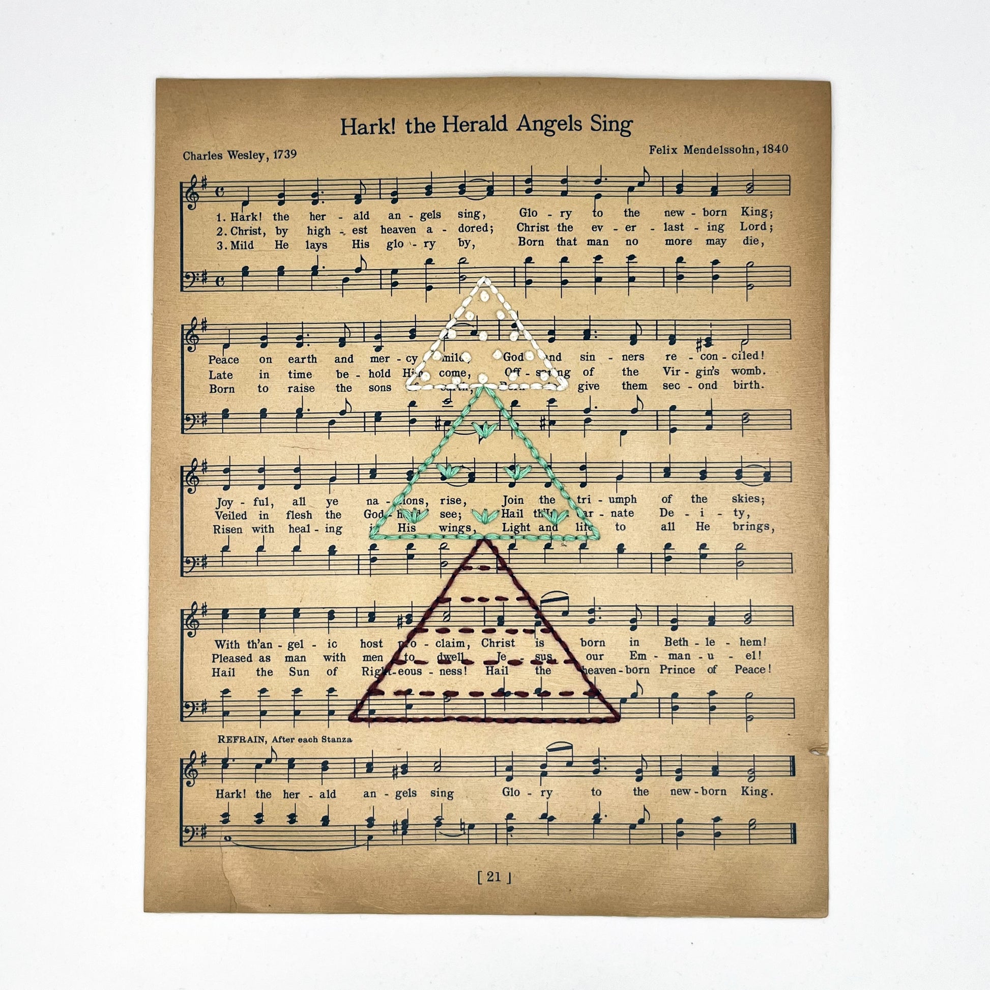 Christmas sheet music for the song Hark the Herald Angels Sing, hand stitched on with a Christmas tree made from triangles filled with different types of stitches, in red, green and ivory thread, on a white background