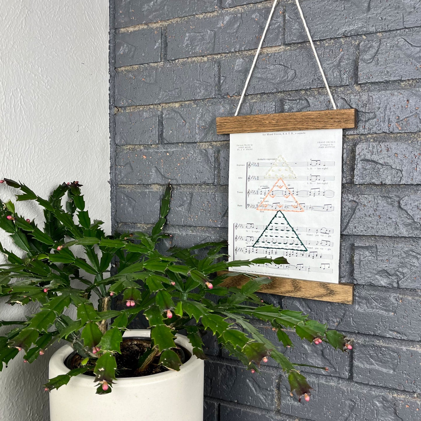 Christmas sheet music, hand stitched on with a Christmas tree made from triangles filled with different types of stitches, in green, peach and ivory thread, hanging in a wood magnetic frame on a grey brick wall, with a Christmas cactus in front of it