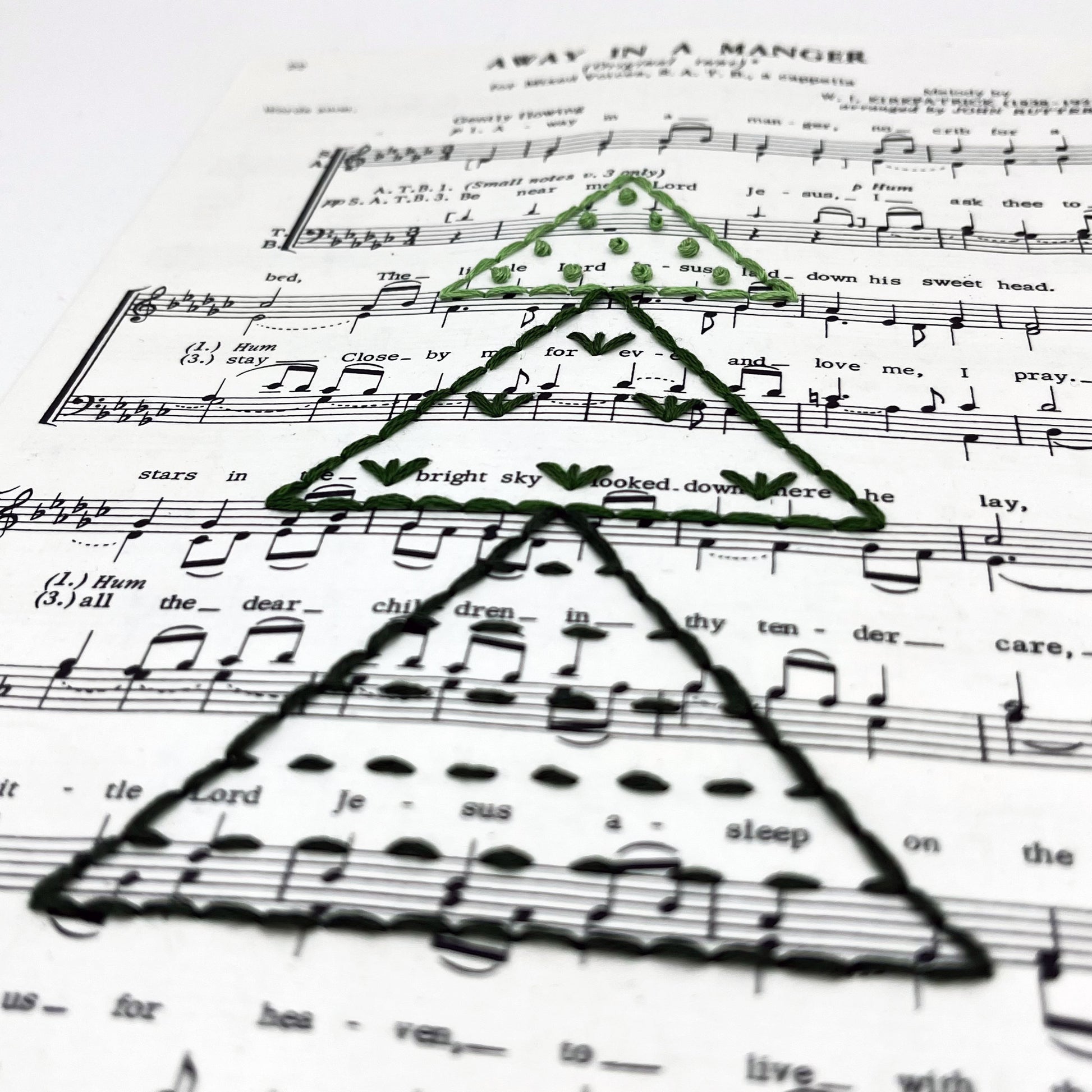 close up angled view of Christmas sheet music for the song Away in a Manger, hand stitched on with a Christmas tree made from triangles filled with different types of stitches, in shades of green thread