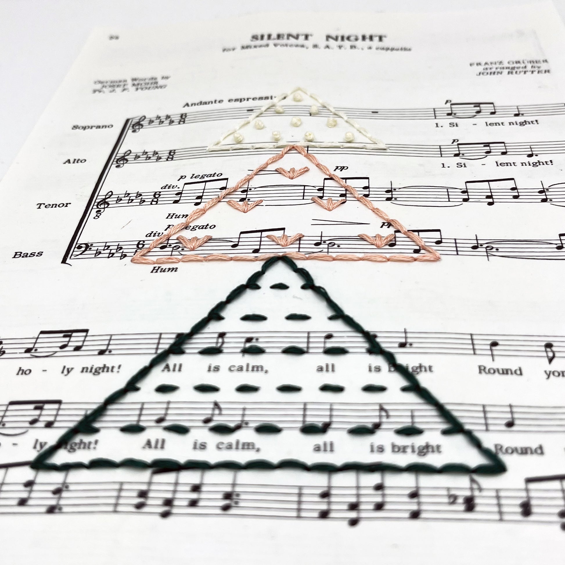 close up angled view of Christmas sheet music for the song Silent Night, hand stitched on with a Christmas tree made from triangles filled with different types of stitches, in green, peach and ivory thread