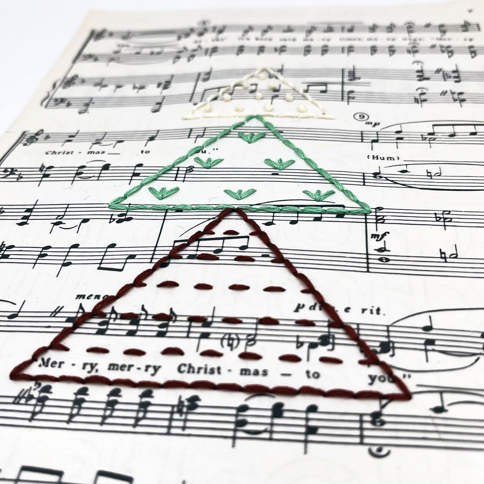 close up angled view of Christmas sheet music for the song The Christmas Song, hand stitched on with a Christmas tree made from triangles filled with different types of stitches, in red, green and ivory thread