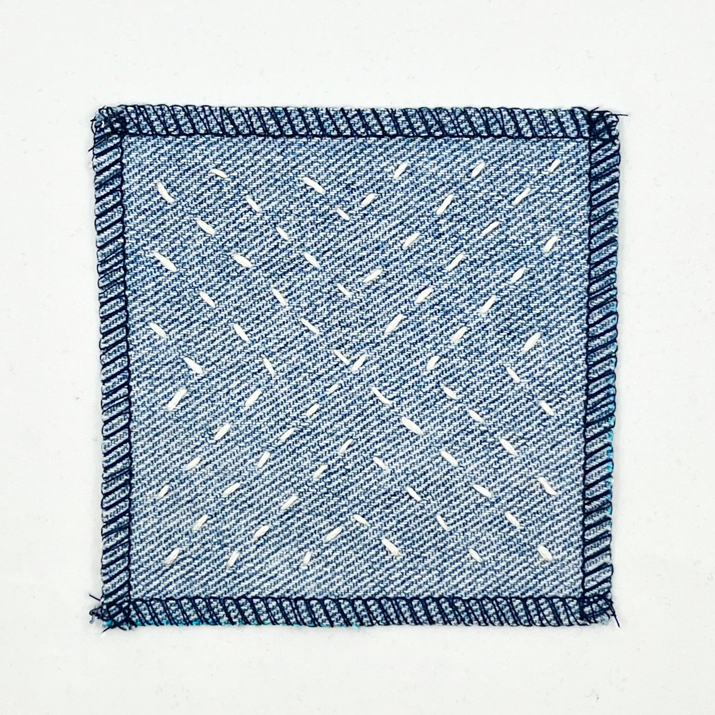 a square patch made out of denim, handstitched with ivory running stitches in a radiating X pattern, with overlocked edges, on a white background
