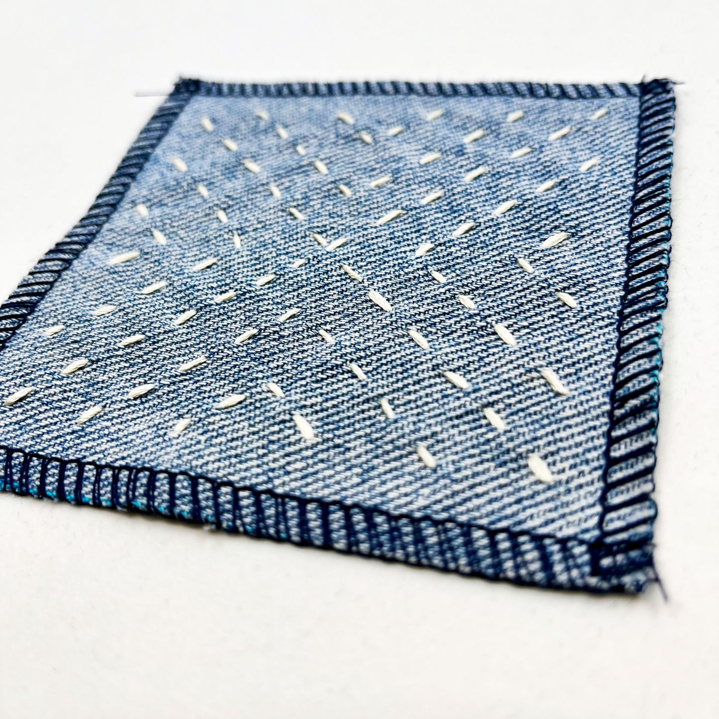close up angled view of a square patch made out of denim, handstitched with ivory running stitches in a radiating X pattern, with overlocked edges, on a white background