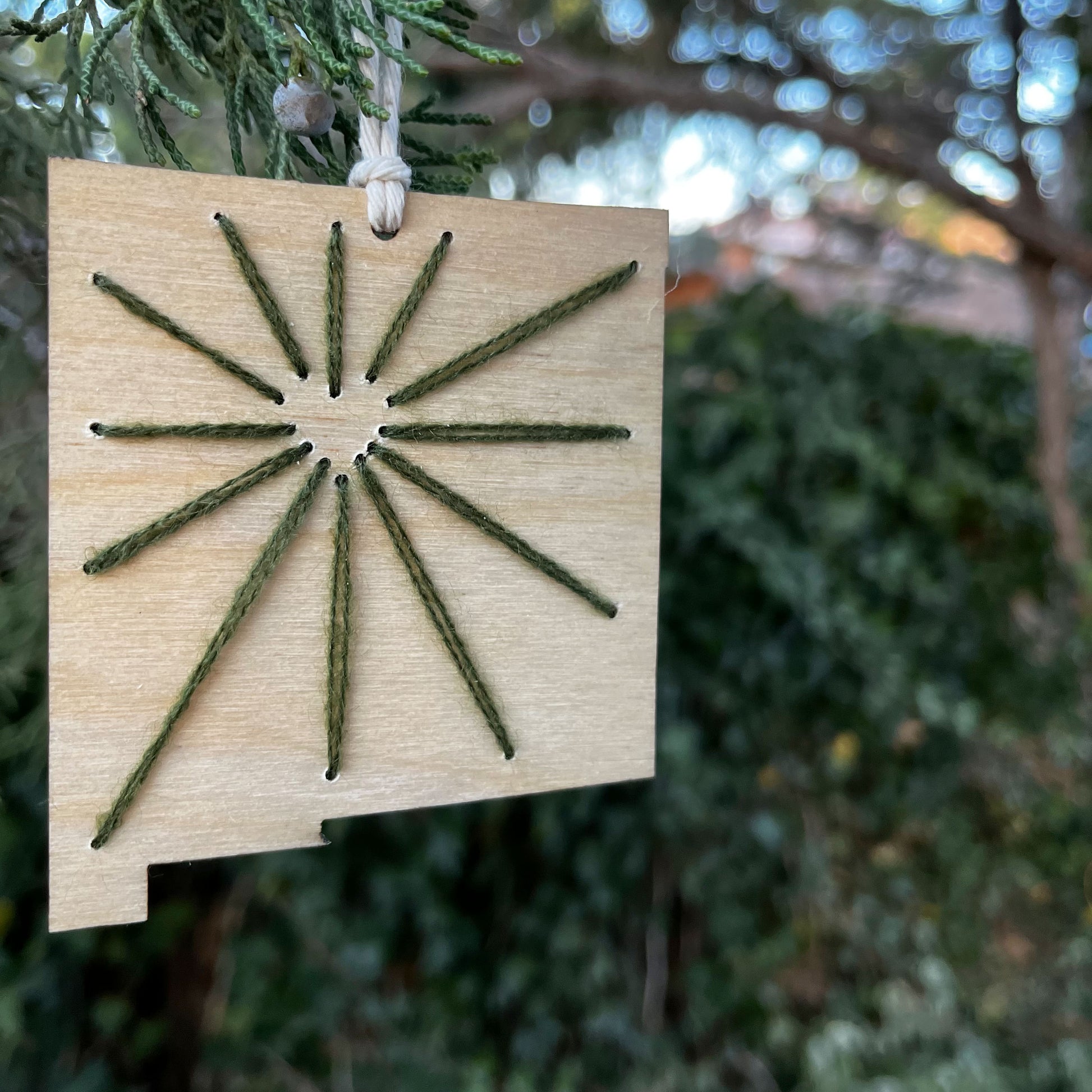 a laser cut wood ornament in the shape of New Mexico, hand embroidered in pine green with negative space in the shape of a heart where Albuquerque is, and lines coming out from it like sunbeams, with an off white cord loop hanging on a pine tree