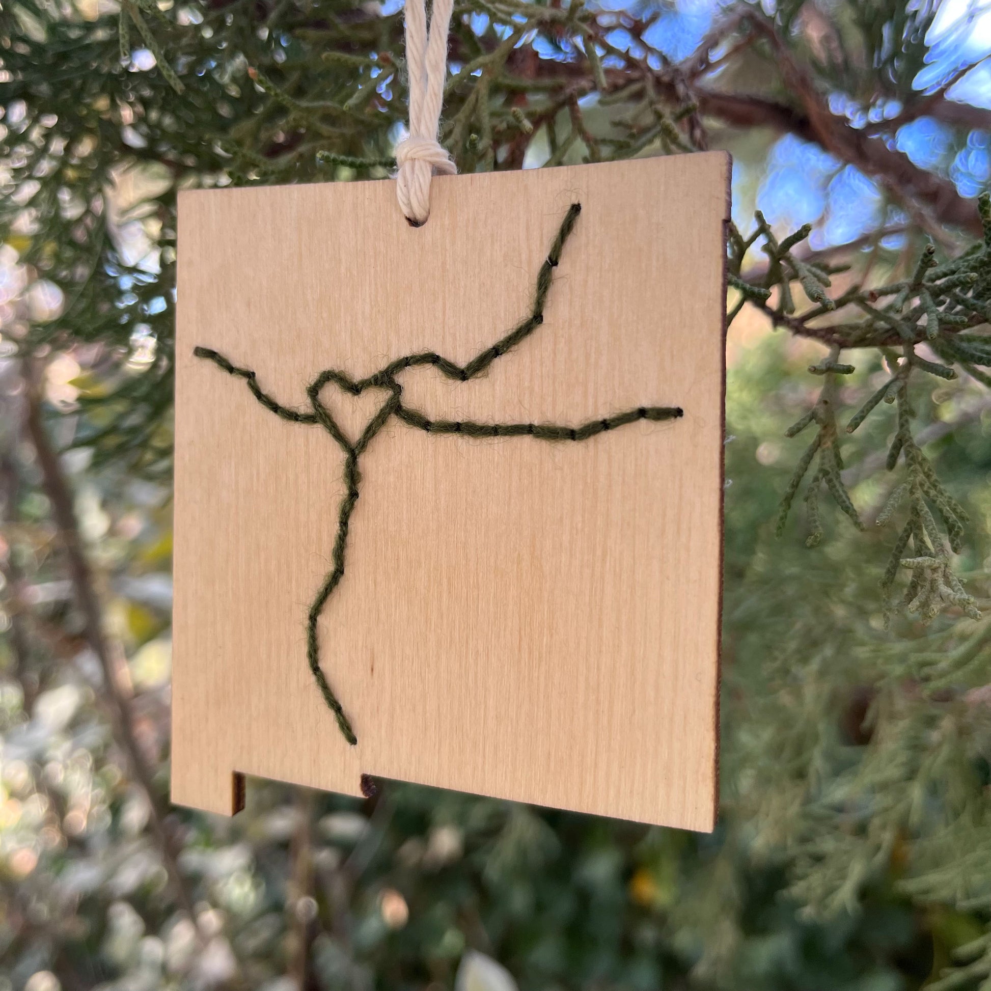 a laser cut wood ornament in the shape of New Mexico, hand embroidered in pine green with a heart where Albuquerque is, and Interstate 25 and 40, with an off white cord loop hanging on a pine tree